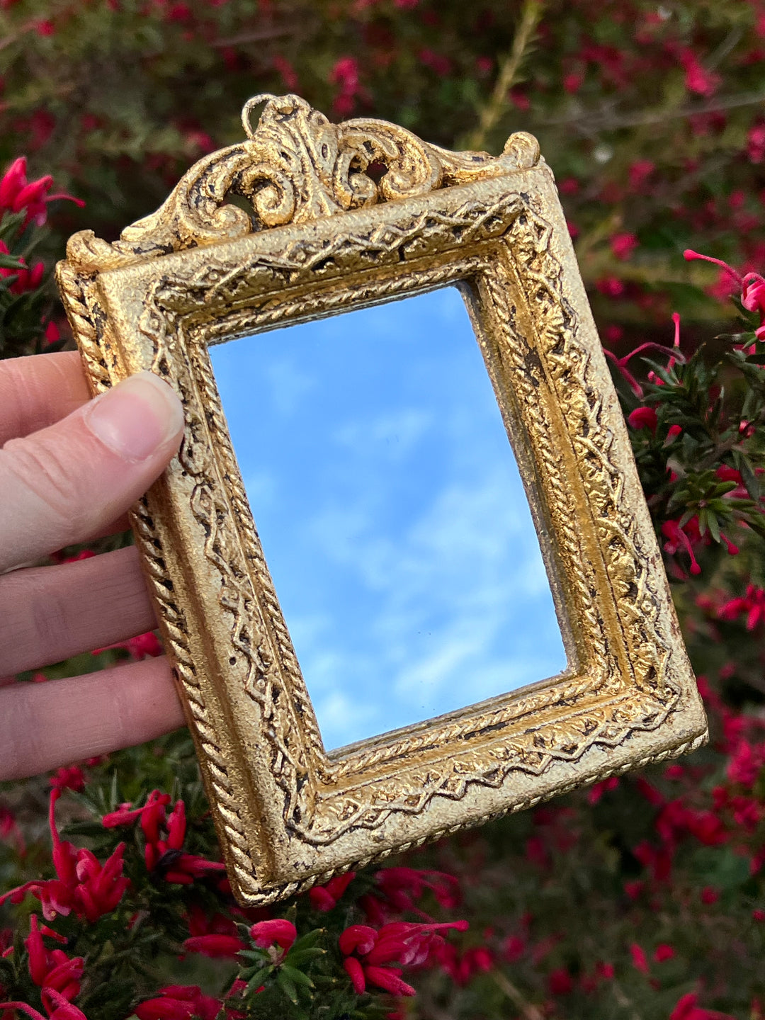 Specchietto di misure 14x10 cm, di ispirazione barocca con cornice intagliata e dorata a foglia oro. Nella parte superiore è presente un dettaglio a fregio e lungo la cornice un decoro geometrico. Con una maestria che trascende il tempo, i nostri specchietti sono capolavori realizzati a mano che catturano l'essenza dell'eleganza barocca. Ogni cornice, finemente scolpita e rifinita con foglia d'oro, è frutto di tecniche di antichizzazione che regalano un'anima vissuta e senza tempo ad ogni pezzo.